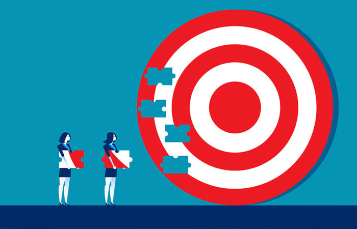 Two businesswomen carrying puzzle pieces towards a large jigsaw of a bullseye target. Illustration.